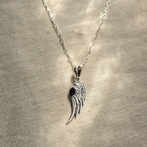 K&ouml;nnte beinhalten: Silberfarbene Halskette mit Anh&auml;nger in Form eines detaillierten Fl&uuml;gels mit einem dunkelblauen Stein. Der Fl&uuml;gelanh&auml;nger h&auml;ngt an einer filigranen Kette vor einem neutralen Stoffhintergrund.