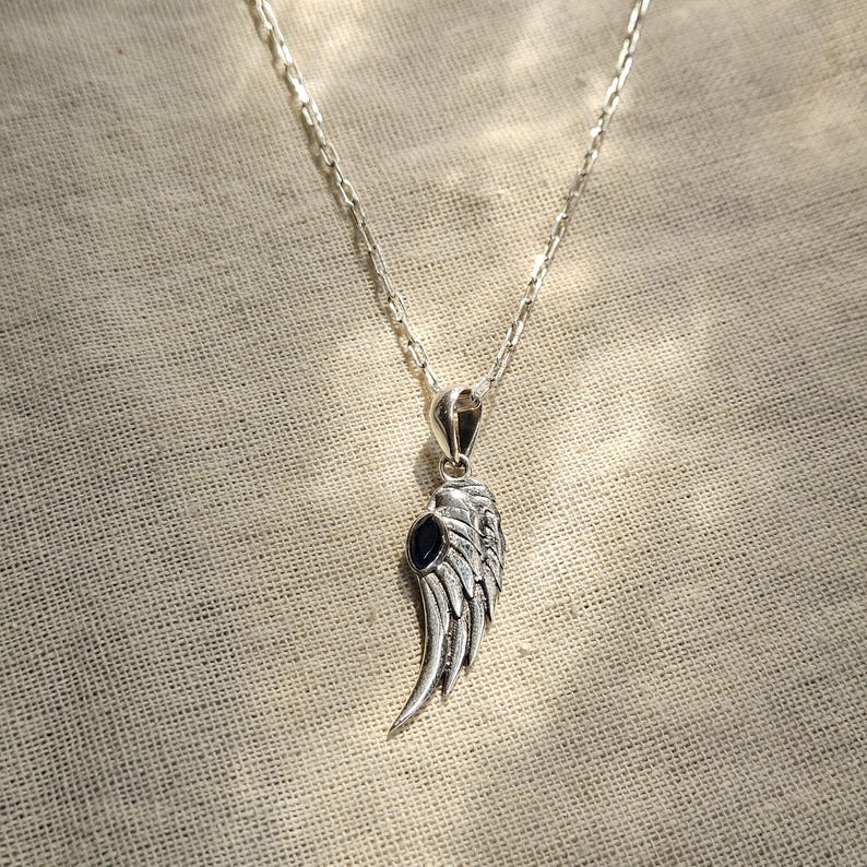 K&ouml;nnte beinhalten: Silberfarbene Halskette mit Anh&auml;nger in Form eines detaillierten Fl&uuml;gels mit einem dunkelblauen Stein. Der Fl&uuml;gelanh&auml;nger h&auml;ngt an einer filigranen Kette vor einem neutralen Stoffhintergrund.