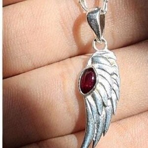 K&ouml;nnte beinhalten: Halskette mit einem silberfarbenen Anh&auml;nger in Fl&uuml;gelform mit einem roten Edelstein. Der Fl&uuml;gel hat detaillierte Federstrukturen und h&auml;ngt an einer feinen Kette. Der Edelstein ist oval. Schmuck.