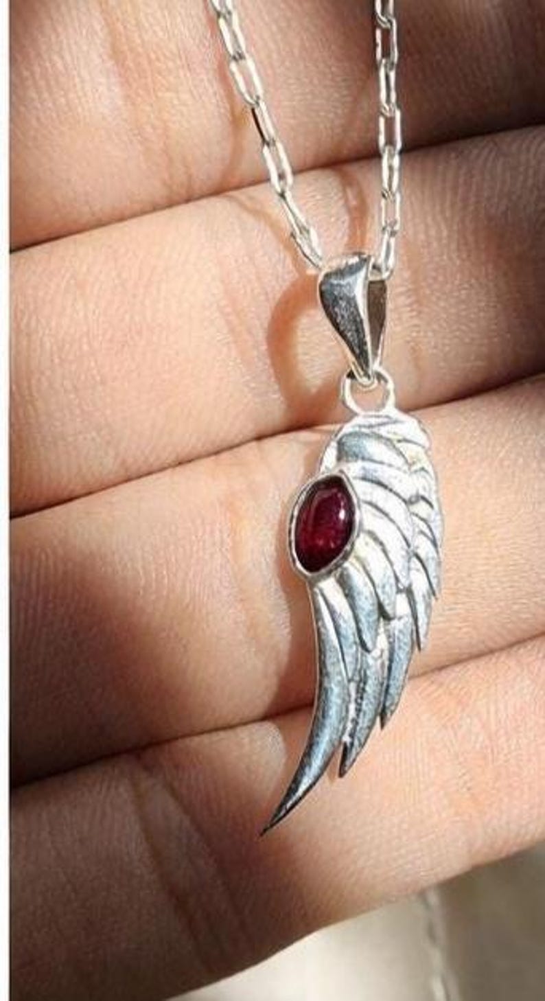 K&ouml;nnte beinhalten: Halskette mit einem silberfarbenen Anh&auml;nger in Fl&uuml;gelform mit einem roten Edelstein. Der Fl&uuml;gel hat detaillierte Federstrukturen und h&auml;ngt an einer feinen Kette. Der Edelstein ist oval. Schmuck.