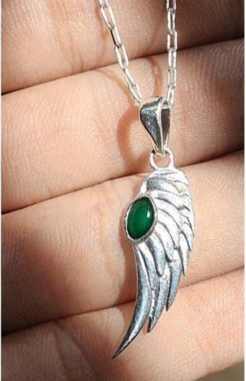 K&ouml;nnte beinhalten: Eine silberfarbene Halskette mit einem Anh&auml;nger in Fl&uuml;gelform und einem gr&uuml;nen Edelstein. Der Fl&uuml;gelanh&auml;nger ist mit federartigen Texturen versehen und h&auml;ngt an einer filigranen Kette. Der gr&uuml;ne Edelstein ist oval und in der Mitte des Fl&uuml;gels gefasst.