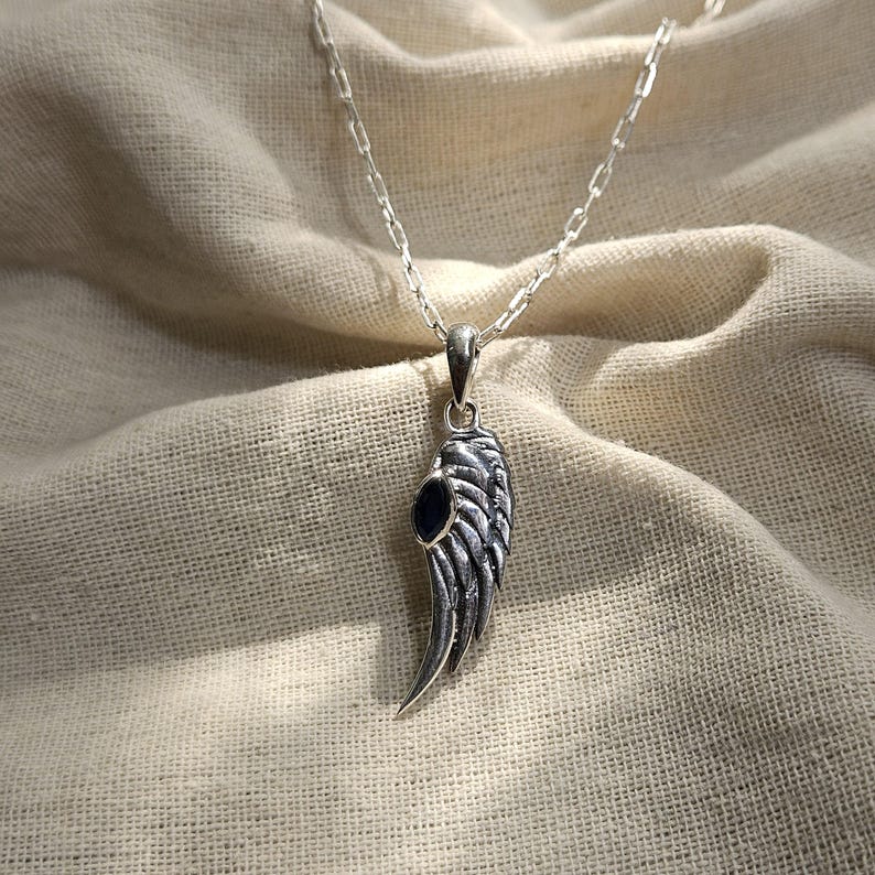 K&ouml;nnte beinhalten: Halskette mit silberfarbenem Anh&auml;nger in Form eines Fl&uuml;gels. Der Anh&auml;nger hat ein detailliertes Fl&uuml;geldesign mit einem dunklen, oval geformten Stein. Die zarte Kette bildet einen Kontrast zum neutralen Hintergrund.