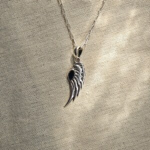 K&ouml;nnte beinhalten: Halskette mit silberfarbenem Anh&auml;nger in Fl&uuml;gelform und einem dunkelblauen Stein. Der Fl&uuml;gelanh&auml;nger mit federartigen Details h&auml;ngt an einer feinen Kette. Die Halskette wird vor einem strukturierten, beigefarbenen Stoffhintergrund gezeigt.