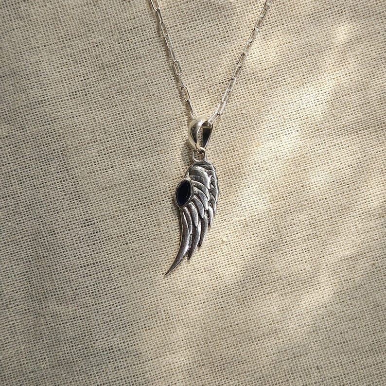 K&ouml;nnte beinhalten: Halskette mit silberfarbenem Anh&auml;nger in Fl&uuml;gelform und einem dunkelblauen Stein. Der Fl&uuml;gelanh&auml;nger mit federartigen Details h&auml;ngt an einer feinen Kette. Die Halskette wird vor einem strukturierten, beigefarbenen Stoffhintergrund gezeigt.