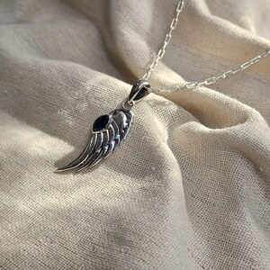 K&ouml;nnte beinhalten: Halskette mit silberfarbenem Anh&auml;nger in Form eines Fl&uuml;gels mit einem dunkelblauen Akzentstein. Der Fl&uuml;gelanh&auml;nger ist an einer filigranen Kette befestigt. Die Halskette wird auf einem neutralfarbenen Stoff pr&auml;sentiert.