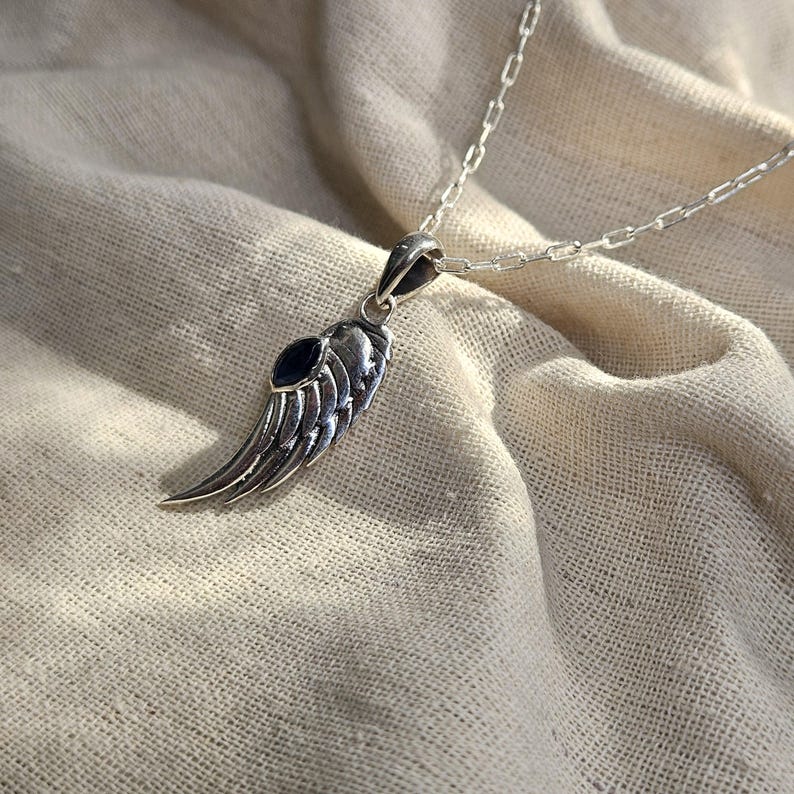 K&ouml;nnte beinhalten: Halskette mit silberfarbenem Anh&auml;nger in Form eines Fl&uuml;gels mit einem dunkelblauen Akzentstein. Der Fl&uuml;gelanh&auml;nger ist an einer filigranen Kette befestigt. Die Halskette wird auf einem neutralfarbenen Stoff pr&auml;sentiert.