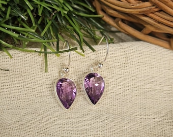 May include: A pair of silver drop earrings featuring pear-shaped, faceted amethyst gemstones. The earrings have a simple design with a small ball detail at the top of the hook. The purple gemstones are set in a silver bezel.