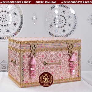 Può includere: Una scatola decorativa rosa con bordi dorati e un motivo floreale. La scatola ha una maniglia in ottone e due nappine. La scatola è decorata con paillettes e una corona dorata con le lettere "SRK" al centro. Il testo "Jalandhar" e "Box" è anche visibile sulla scatola.