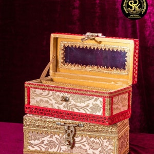 May include: Two stacked ornate boxes with gold trim and a floral pattern. The top box is red with a gold interior and a clear lid. The bottom box is cream with a gold interior. The boxes are on a red velvet surface. The text "SRK Bangles +918360721433 Attari bazar Jalandhar" is visible at the bottom of the image.