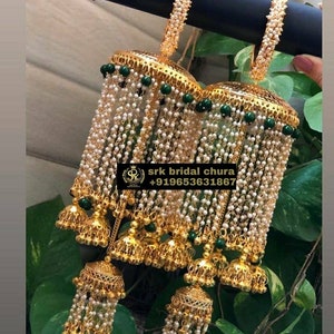 Può includere: Un paio di braccialetti dorati con dettagli di perle bianche e perline verdi. I braccialetti sono collegati a un set di campane dorate con perline verdi e fili di perle bianche.