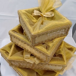 May include: A stack of four square gift boxes. Each box is covered in gold fabric and trimmed with gold decorative trim. The top box is tied with a gold bow. The boxes are arranged in a pyramid shape.