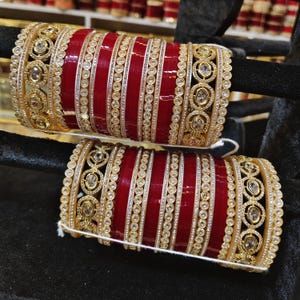 Può includere: Un set di braccialetti rossi e dorati con pietre bianche. I braccialetti sono impilati uno sopra l'altro e sono decorati con disegni intricati.