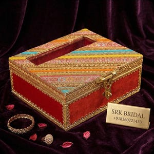 Puede incluir: Una caja rectangular roja con una tapa decorativa con un patrón de rayas de colores. La caja tiene adornos dorados y un cierre de broche. Una pulsera y un anillo dorados están al lado de la caja. El texto "SRK BRIDAL" está en una placa dorada.