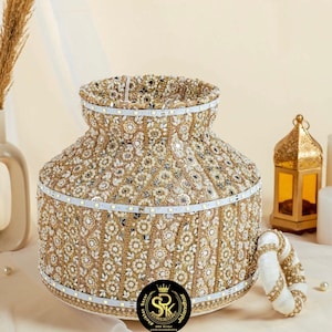 Può includere: Un vaso decorativo dorato e bianco con intricate perline e lavorazioni a specchio. Il vaso è decorato con un nastro bianco e un piccolo logo a corona. Il testo "SRK Bridal" è visibile sul logo.