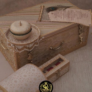 May include: Two ornate boxes with intricate gold and white patterns. The larger box has a scroll with a peacock feather on top. The smaller box has a clear lid revealing a red and gold bangle inside.