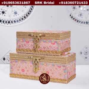 May include: Two pink and gold decorative boxes with intricate floral patterns and ornate brass clasps. The boxes are stacked on top of each other. The top box has a gold trim and the bottom box has a gold trim and a gold logo with the letters "SK" in a crown.