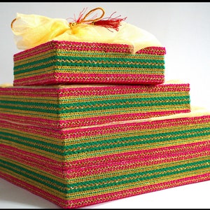 May include: A tiered set of decorative boxes in varying sizes, stacked to resemble a pyramid. The boxes are adorned with alternating bands of green, gold, and pink trim. A yellow fabric with a tassel tops the uppermost box, adding a touch of elegance.