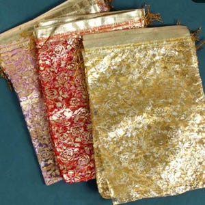 May include: A collection of decorative gift bags in various colors. The bags are gold, red, and lavender, with a floral pattern. Each bag has a gold drawstring closure and trim.