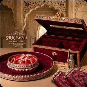 Può includere: Un portagioie in velluto bordeaux con finiture dorate e una chiusura decorativa, aperto per rivelare un interno in peluche. Un vassoio e una ciotola abbinati contengono braccialetti. Sono presenti anche un piccolo flacone di profumo e una collana di perle.