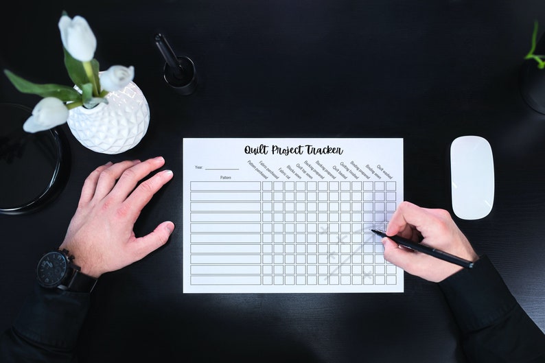 May include: A black and white printable quilt project tracker with columns for year, pattern, fabric yardage, cutting time, quilting time, binding time, and quilt finished. A person is checking off a box on the tracker with a black pen.
