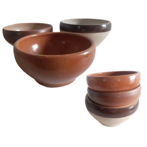 May include: Set of four terracotta bowls, three stacked on top of each other. The top bowl has a brown rim, the middle bowl is plain terracotta, and the bottom bowl has a brown rim and a white base.