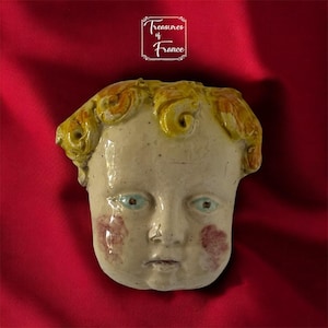 Könnte beinhalten: Keramikskulptur eines Kindergesichts mit gelbem Haar und blauen Augen. Das Gesicht hat rote Wangenröte und befindet sich vor einem roten Hintergrund. Der Text "Treasures of France" befindet sich oben rechts.