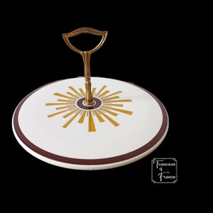 Könnte beinhalten: Ein weißes Keramik-Serviertablett mit einem goldenen Sonnenstrahlen-Design und einem kastanienbraunen Rand. Das Tablett hat einen Messinggriff.