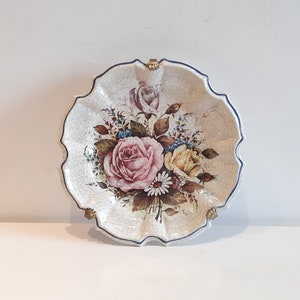 Pode incluir: Um prato de parede de cerâmica branca com uma borda azul e detalhes dourados. O prato apresenta um design floral com rosas cor-de-rosa, amarelas e brancas.