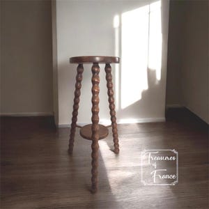 May include: A dark brown wooden plant stand with a round top and a lower shelf. The stand has three turned legs with a bobbin design. The stand is approximately 30 inches tall.