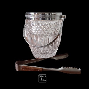 Puede incluir: Un cubo de hielo de vidrio transparente con un borde y un asa plateados, y un par de pinzas plateadas con un mango de madera.