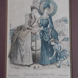 Puede incluir: Una ilustraci&oacute;n vintage de la revista Journal des Demoiselles, julio de 1887, que presenta a tres mujeres con ropa de moda. Dos mujeres est&aacute;n de pie en una playa, una sosteniendo un paraguas, mientras que una ni&ntilde;a peque&ntilde;a con un vestido a rayas juega en la arena. La ilustraci&oacute;n est&aacute; realizada en un estilo delicado, pintado a mano, con colores suaves.