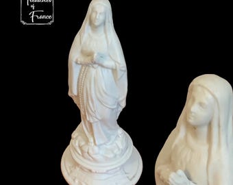 Puede incluir: Dos estatuas de porcelana blanca de la Virgen María. Ambas estatuas están de pie y llevan una túnica larga. La estatua de la izquierda sostiene un rosario en su mano derecha y una flor en su mano izquierda. La estatua de la derecha tiene las manos juntas en oración.