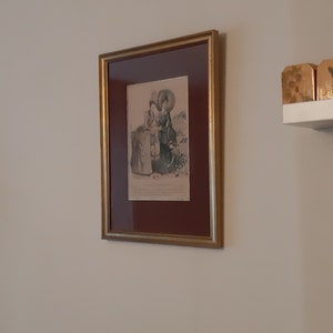 Puede incluir: Una impresi&oacute;n enmarcada de una ilustraci&oacute;n vintage que representa a tres mujeres con vestidos largos y sombreros, de pie al aire libre. La impresi&oacute;n est&aacute; en un marco dorado con un passe-partout marr&oacute;n.