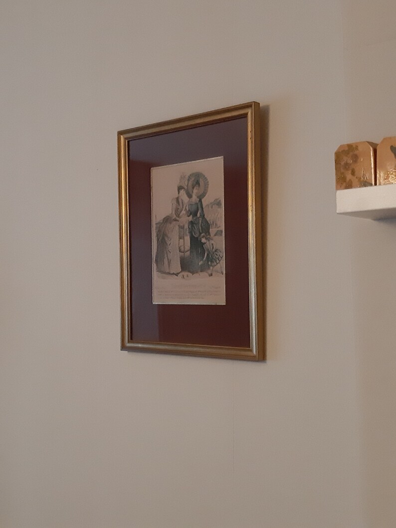 Puede incluir: Una impresi&oacute;n enmarcada de una ilustraci&oacute;n vintage que representa a tres mujeres con vestidos largos y sombreros, de pie al aire libre. La impresi&oacute;n est&aacute; en un marco dorado con un passe-partout marr&oacute;n.