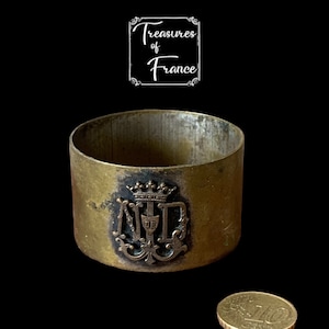 Puede incluir: Un anillo de servilleta de metal dorado con un escudo negro y dorado que presenta las letras "N.D." y una corona. El anillo tiene un diámetro de aproximadamente 5 cm.