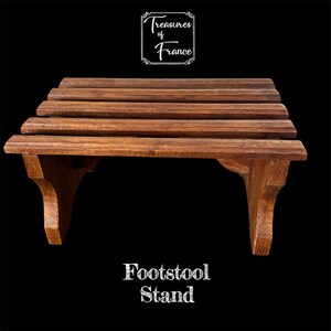 May include: A wooden footstool stand with a dark brown finish. The stand has a slatted top and decorative supports. The text "Treasures of France" is at the top, and "Footstool Stand" is at the bottom.