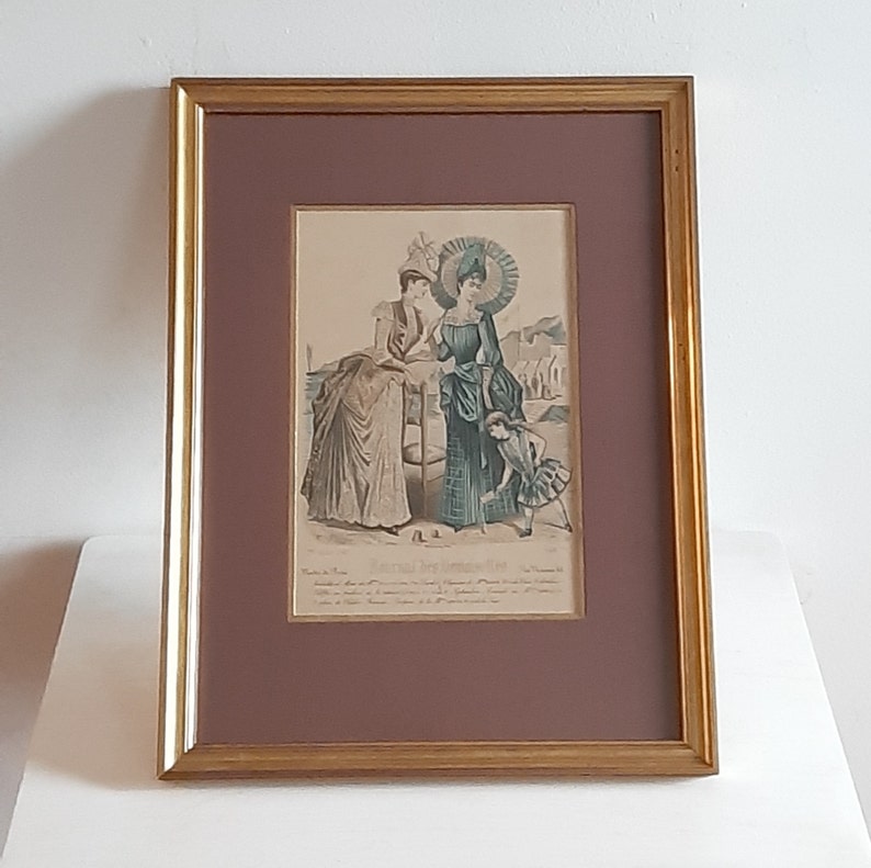 Puede incluir: Una ilustraci&oacute;n vintage enmarcada de tres mujeres con ropa de moda de finales del siglo XIX. Las mujeres est&aacute;n de pie en una playa con vista al mar y un muelle en el fondo. La ilustraci&oacute;n est&aacute; en tonos azules, verdes y marrones. El texto en la parte inferior dice "Modes et Fantaisies."