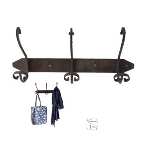 May include: A rustic, black wrought iron coat rack with three hooks. The rack is mounted on a wooden board and has a decorative, pointed end. A blue tote bag and a blue scarf are hanging on the hooks.