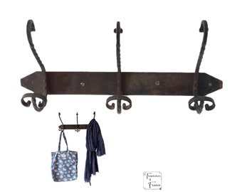 Op de afbeelding: Een rustieke, zwarte smeedijzeren kapstok met drie haken. De kapstok is gemonteerd op een houten plank en heeft een decoratief, puntig uiteinde. Een blauwe boodschappentas en een blauwe sjaal hangen aan de haken.