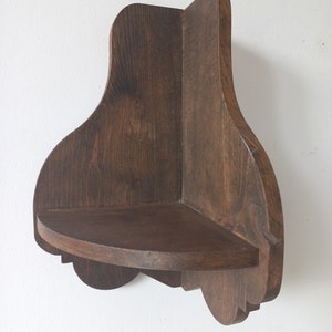 K&ouml;nnte beinhalten: Ein dunkelbraunes Eckregal aus Holz mit einer abgerundeten Vorderkante und dekorativen Details. Das Regal ist an einer wei&szlig;en Wand montiert.