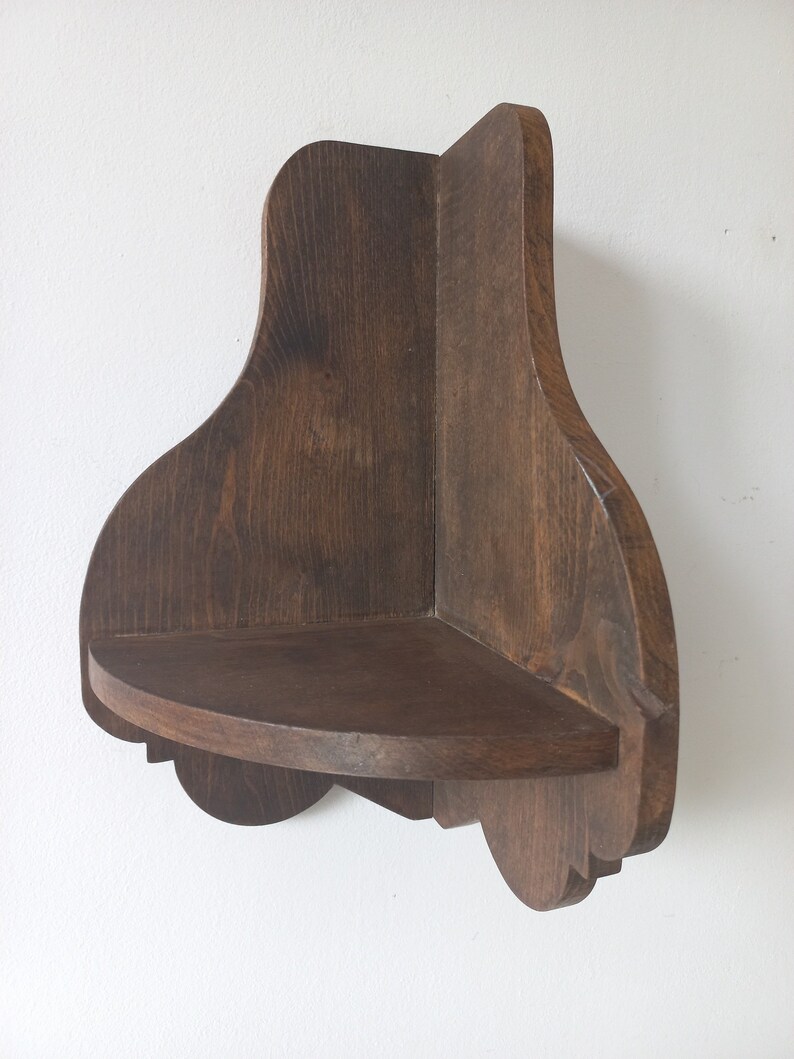 K&ouml;nnte beinhalten: Ein dunkelbraunes Eckregal aus Holz mit einer abgerundeten Vorderkante und dekorativen Details. Das Regal ist an einer wei&szlig;en Wand montiert.