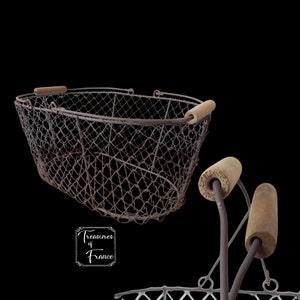 Puede incluir: Una cesta de alambre vintage con asas de madera. La cesta es de forma ovalada y está hecha de malla de alambre. La cesta está oxidada y tiene un aspecto desgastado. La cesta está sobre un fondo negro.