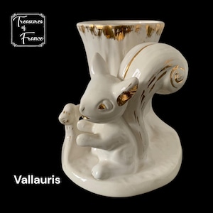 Könnte beinhalten: Weiße Keramik-Eichhörnchenfigur mit goldenen Akzenten. Das Eichhörnchen sitzt auf einem Baumstamm und hält eine Nuss in seiner Pfote. Die Figur ist eine Vase mit einem goldenen Rand.