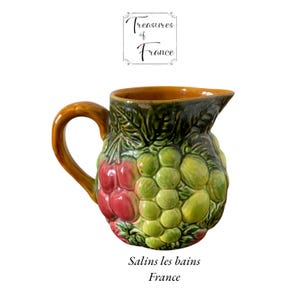 Könnte beinhalten: Keramikkrug mit Henkel, verziert mit einem Traubenmuster in Rot, Grün und Gelb. Der Krug hat einen braunen Henkel und Rand, mit dem Text "Treasures of France" und "Salins les bains France".