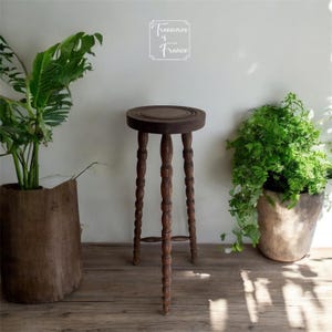 May include: A dark brown, antique wooden plant stand with a round top and three turned legs. The stand is approximately 28 inches tall. The stand is surrounded by plants in decorative pots, creating a natural, rustic aesthetic.