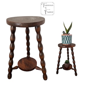 May include: Two dark brown wooden stools. The larger stool has a round top and a lower shelf. The legs are turned with a bobbin design. The smaller stool has a plant on top and a cactus on the lower shelf. The text "Treasures of France" is in the upper right corner.