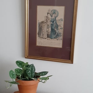 Puede incluir: Una ilustraci&oacute;n vintage enmarcada de dos mujeres con ropa de moda de finales del siglo XIX. Las mujeres est&aacute;n de pie en una playa con un ni&ntilde;o peque&ntilde;o delante de ellas. La ilustraci&oacute;n est&aacute; en un marco dorado con un paspart&uacute; marr&oacute;n.