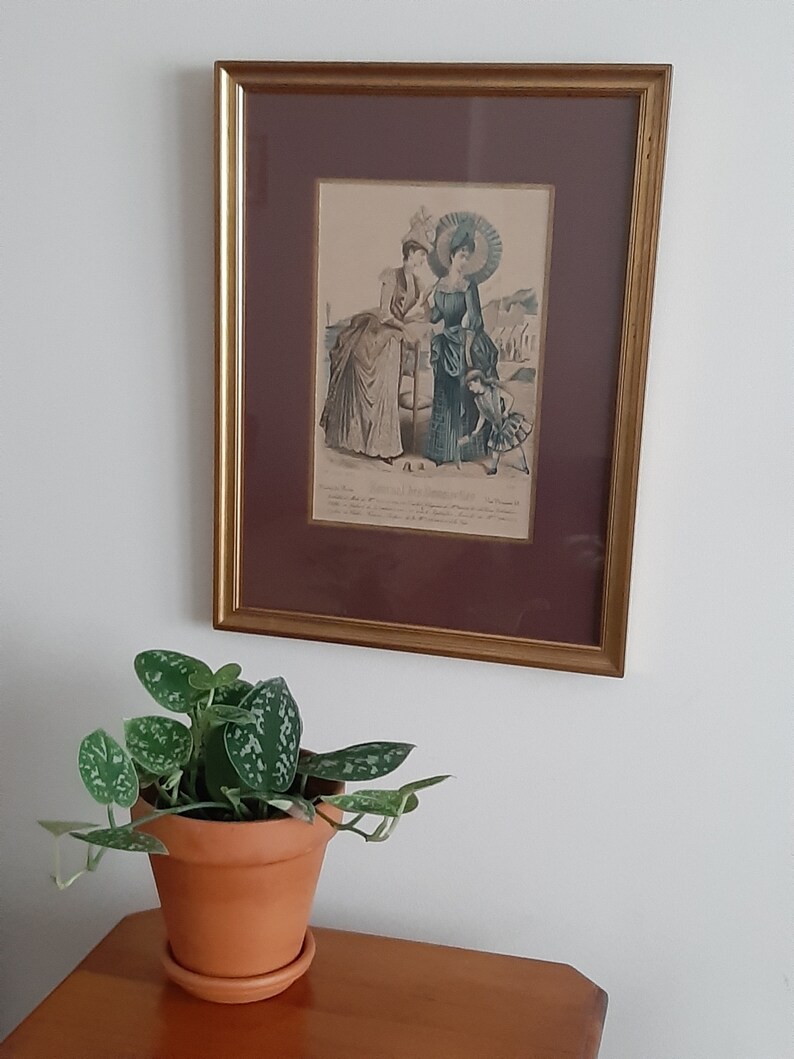 Puede incluir: Una ilustraci&oacute;n vintage enmarcada de dos mujeres con ropa de moda de finales del siglo XIX. Las mujeres est&aacute;n de pie en una playa con un ni&ntilde;o peque&ntilde;o delante de ellas. La ilustraci&oacute;n est&aacute; en un marco dorado con un paspart&uacute; marr&oacute;n.
