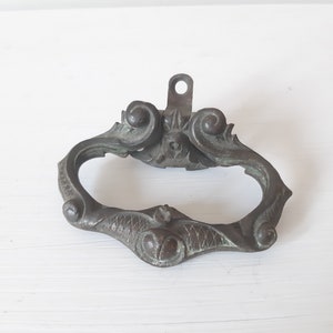 Op de afbeelding: Een antieke bronzen deurgreep met sierlijke krulwerk en een ringvormige handgreep. De greep heeft een verweerde afwerking en is ontworpen in een traditionele stijl.