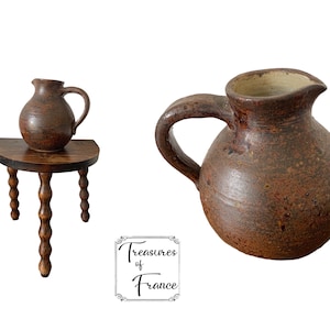 May include: A brown ceramic pitcher with a rounded body and a handle. The pitcher has a speckled brown glaze and is sitting on a wooden table with three legs.